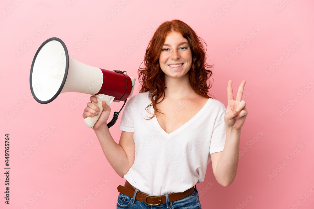 Teenager reddish woman isolated on pink background holding a megaphone and smiling and showing victory sign Teenager reddish woman isolated on pink background holding a megaphone and smiling and showing victory sign