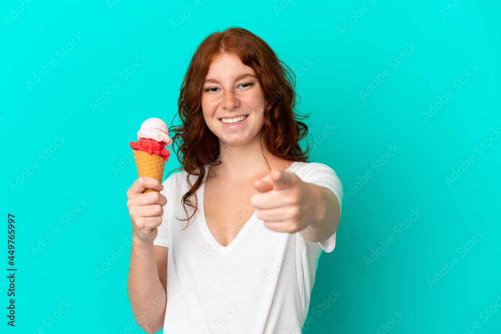 Teenager reddish woman with a cornet ice cream isolated on blue background surprised and pointing front Teenager reddish woman with a cornet ice cream isolated on blue background surprised and pointing front
