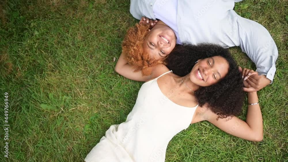 Young beautiful happy lesbian African American couple lying on grass laughing stretching out hands closing from camera, summer top view. LGBT community concept. Female friends enjoying love moments vídeo do Stock | Adobe Stock