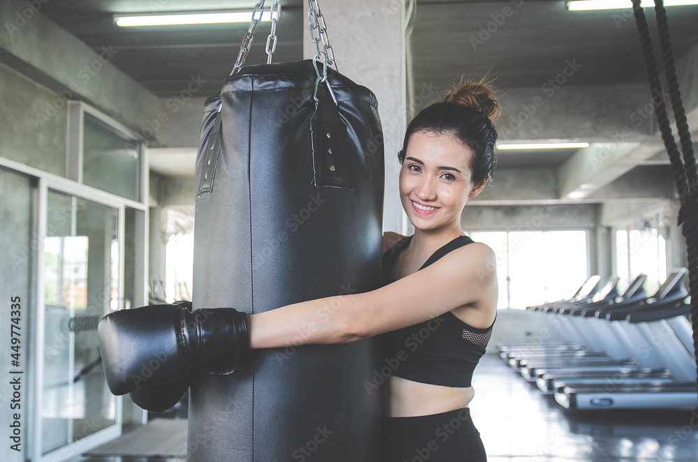 Beautiful asian woman wears boxing gloves hugging a sandbag and smile in gym or sport center