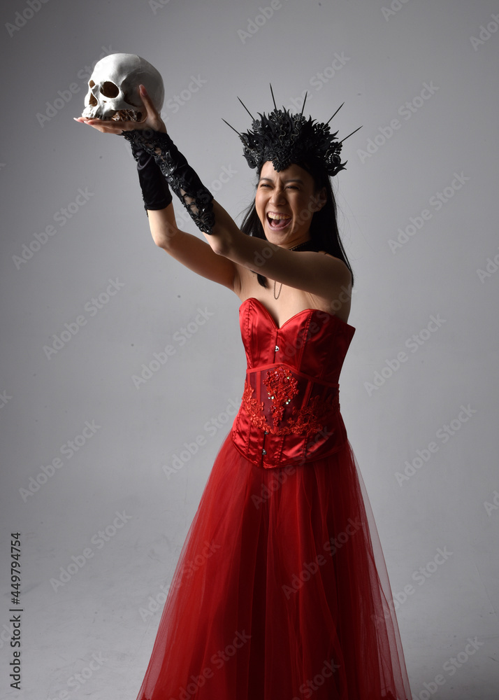 portrait of beautiful young asian woman wearing red corset and crown, posing in gothic horror style with creative hand gestures isolated against dark black studio background. portrait of beautiful young asian woman wearing red corset and crown, posing in gothic horror style with creative hand gestures isolated against dark black studio background.
