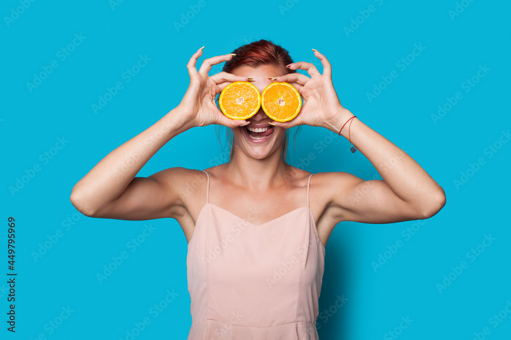 Lovely red haired woman is covering her eye with sliced lemons posing on a blue studio wall