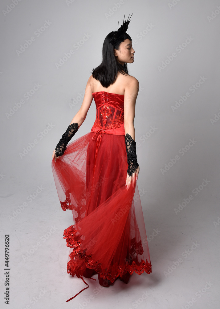 Full length portrait of beautiful young asian woman wearing red corset and ornate gothic queen crown. Graceful standing posing isolated on studio background. Full length portrait of beautiful young asian woman wearing red corset and ornate gothic queen crown. Graceful standing posing isolated on studio background.