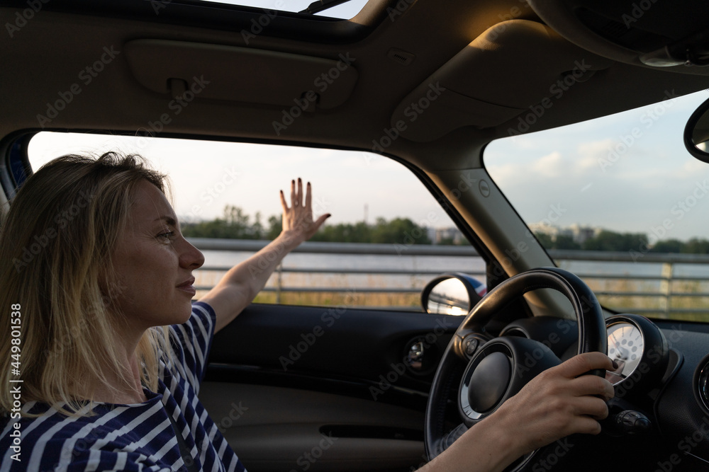 Relaxed calm woman driving car enjoy speed and freedom stretching hand in open window. Confident middle age female driver look at horizon sunset and road. Blonde casual girl in 40s hold steering wheel