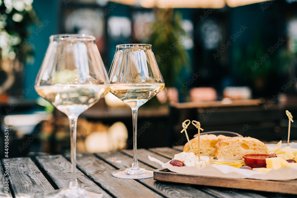 A wooden table in a restaurant with a cheese plate, grape, honey bread and white wine. Wine glasses and cheese served for a party in a blurred bar or a restaurant on terrace at a sunny summer day.