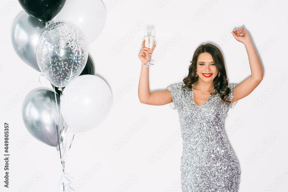 Christmas mood. Happy woman. Festive toast. Holiday celebration. Beautiful smiling lady in spangle silver dress posing with glass of champagne isolated white colorful balloons copy space.