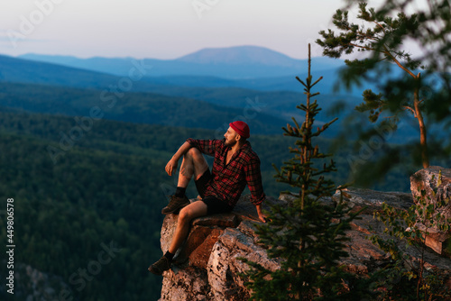 A male traveler is sitting on the edge of a cliff with a view of the forest from a height of flight. Travel as a way of life, adventure, vacation. A traveler at sunset in the mountains. Copy space