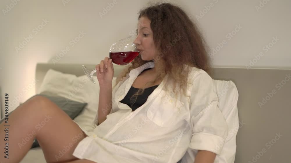 Relaxed young woman in white shirt drinks red wine. Mixed race brunette with bare legs holds wineglass sitting alone on bed. Lonely lady suffers from loneliness at home weekend Relaxed young woman in white shirt drinks red wine. Mixed race brunette with bare legs holds wineglass sitting alone on bed. Lonely lady suffers from loneliness at home weekend