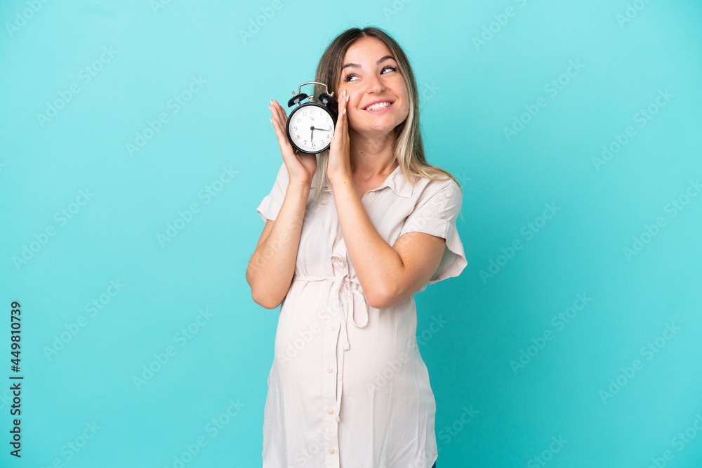 Fototapeta premium Young Romanian woman isolated on blue background pregnant and holding clock
