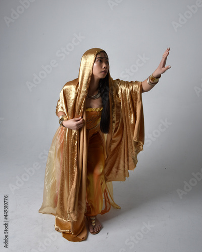 Full length portrait of pretty young asian woman wearing golden Arabian robes like a genie, standing pose holding flowing fabric, isolated on studio background.