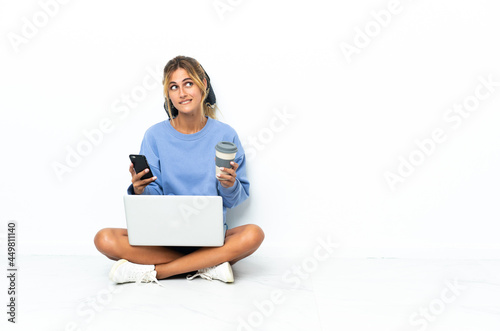Young blonde Uruguayan girl with the laptop isolated on white background holding coffee to take away and a mobile while thinking something