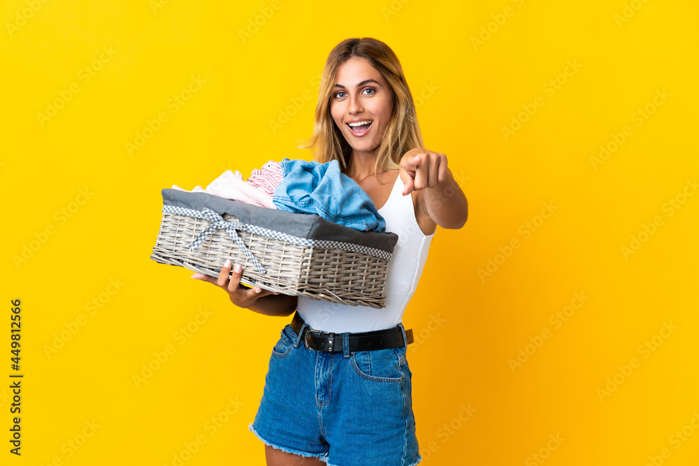 Young blonde Uruguayan woman holding a clothes basket isolated on yellow background surprised and pointing front Young blonde Uruguayan woman holding a clothes basket isolated on yellow background surprised and pointing front