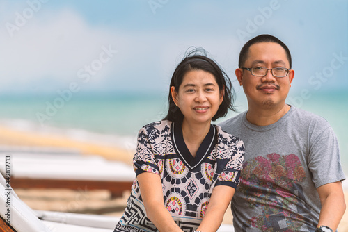 Portrait of Asian middle-aged couple husband and wife sitting on a sunbed on the beach, eyes looking at the camera, blurred background beautiful color of sea and sky. Blank space for copy.