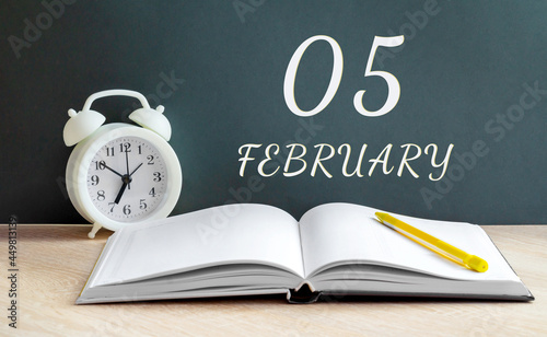 february 05. 05-th day of the month, calendar date.A white alarm clock, an open notebook with blank pages, and a yellow pencil lie on the table.Winter month, day of the year concept photo