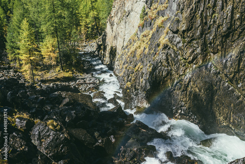 Atmospheric autumn landscape with rainbow above turbulent mountain river among rocks near rocky wall in sunshine. Beautiful alpine scenery with powerful mountain river and autumn forest in sunlight.