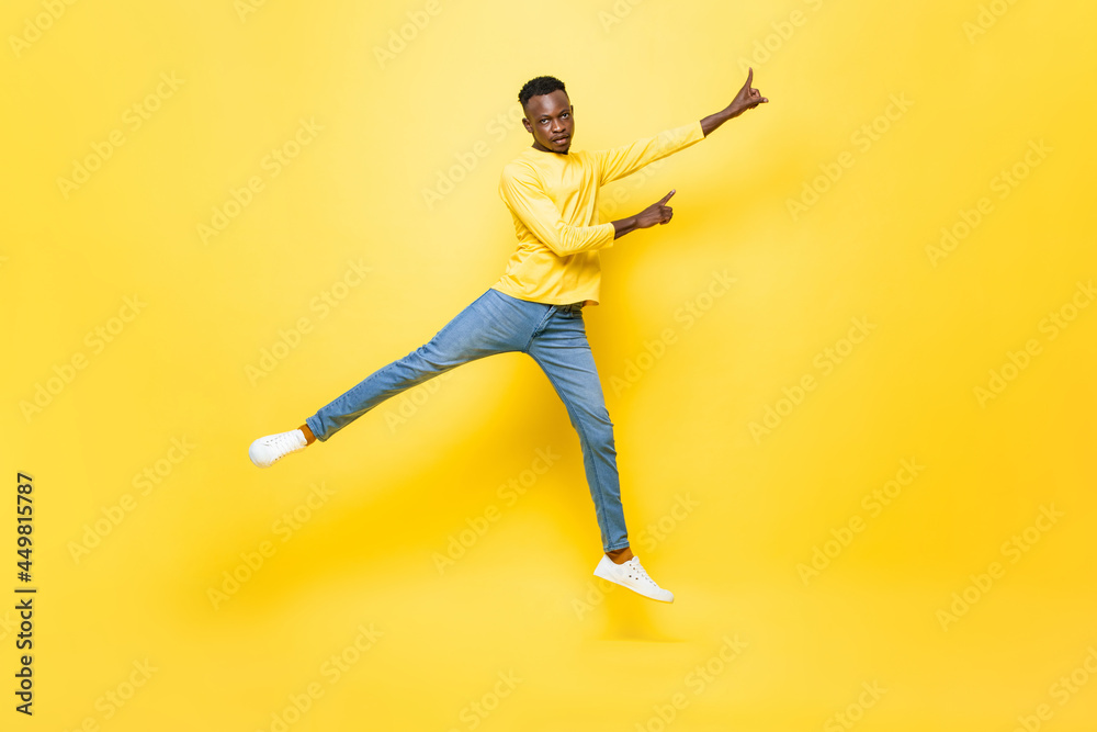 Young energetic African man jumping and pointing hands up on isolated yellow studio background