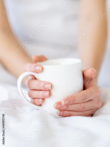 Sunday coffee. Good morning. Home relaxation. Hot beverage. Closeup of elegant woman hands holding cup of latte drink on soft clean white bedsheets blanket in cozy bed.