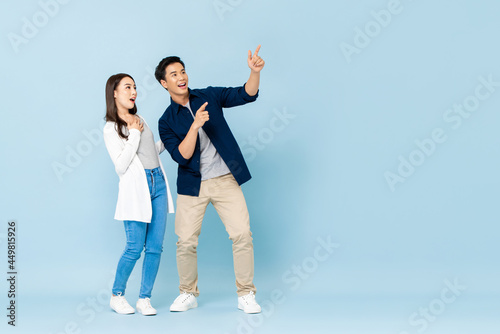 Full length portrait of happy excited Asian couple tourists pointing hands to empty space on isolated light blue background