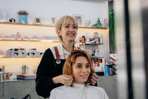 Portrait of happy young Asian woman hairstylist in apron while doing hairdressing and put care treatment while styling curly hair for client