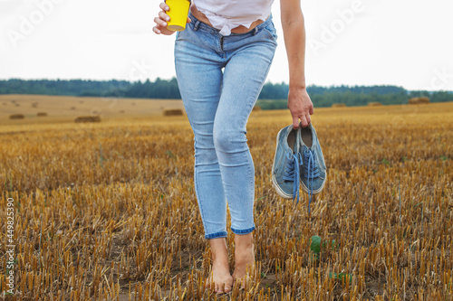 barefoot girl with sneakers and cardboard cup with coffee in hand stand in the agricultural field photo