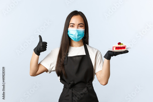 Covid-19, social distancing, small coffee shop business and preventing virus concept. Good-looking smilin gasian female barista, waitress recommend delicious cake in menu, show thumb-up photo