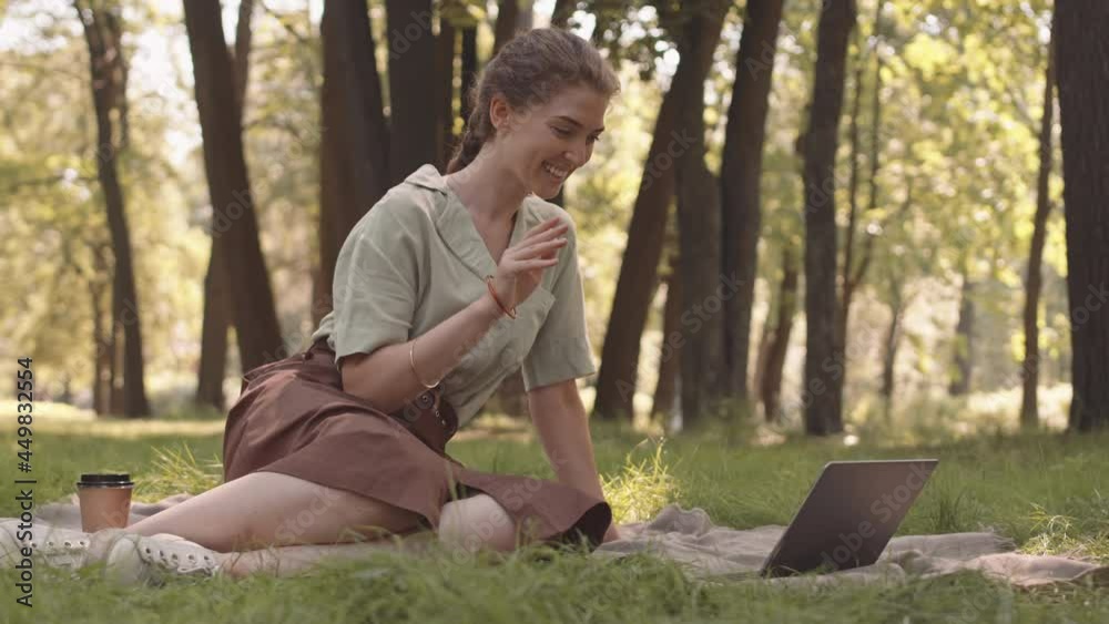 Full shot of young Caucasian woman sitting on blanket on top of lawn in sunny park, video calling using portable computer Full shot of young Caucasian woman sitting on blanket on top of lawn in sunny park, video calling using portable computer