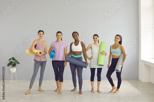 Group portrait of happy beautiful women before or after sports workout. Team of fit young ladies in leggings and yoga pants standing in gym, holding fitness mats, looking at camera and smiling