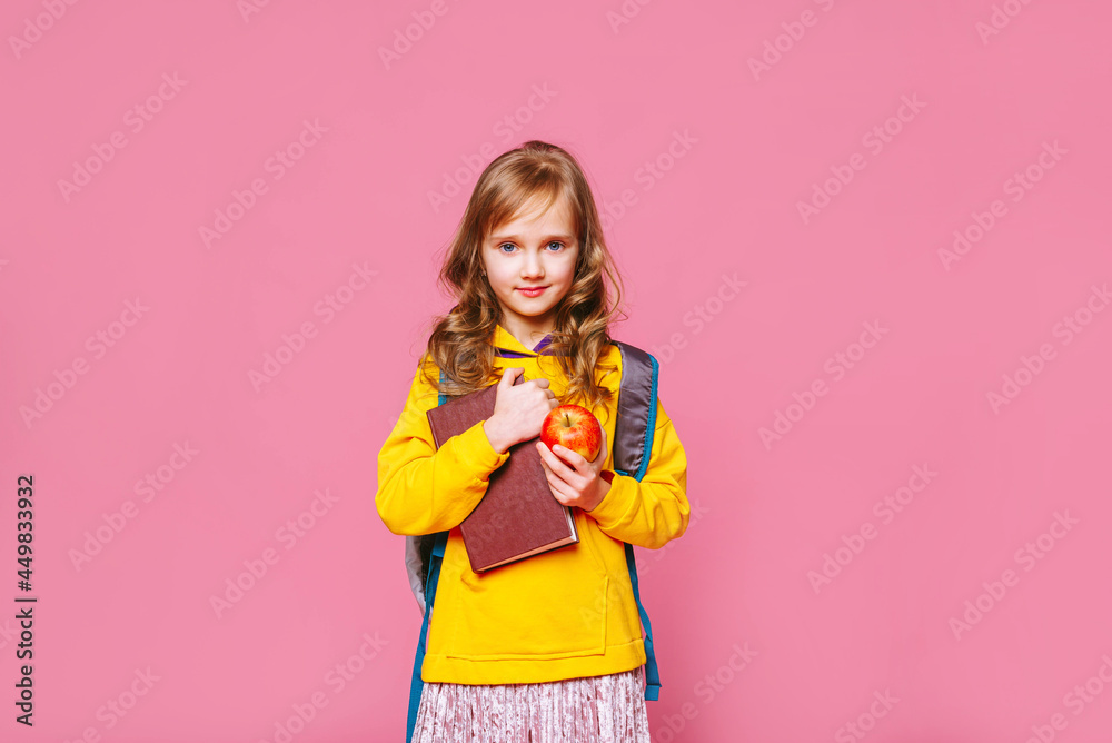 Back to school. Happy blonde girl child  holds a book and a red apple in her hands on a pink background. Education and intellectual development of children. World book day. 1 September.	