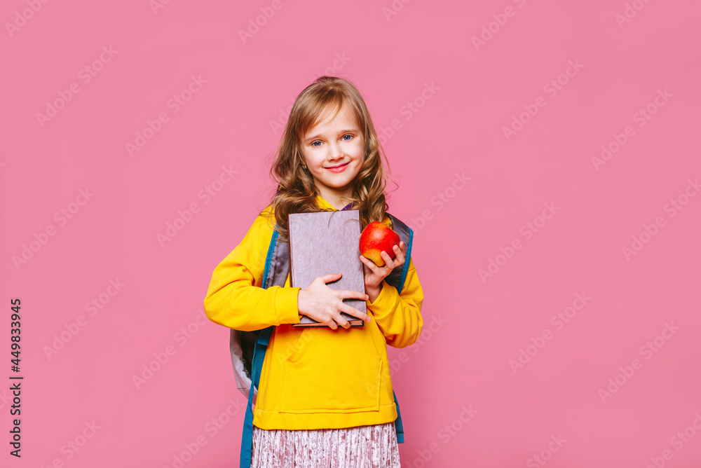 Back to school. Happy blonde girl  child  holds a book,  a red apple in her hand looking at the camera ​on a pink background. Education and intellectual development of children. World book day.