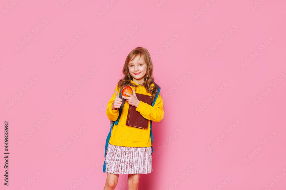 Back to school. Happy blonde girl child holds a book, a red apple in her hand looking at the camera on a pink background. Education and intellectual development of children. World book day. Back to school. Happy blonde girl child holds a book, a red apple in her hand looking at the camera on a pink background. Education and intellectual development of children. World book day.