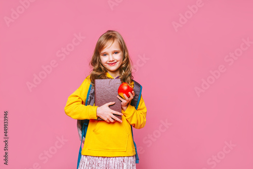 Back to school. Happy blonde girl child holds a book, a red apple in her hand looking at the camera ​on a pink background. Education and intellectual development of children. World book day.