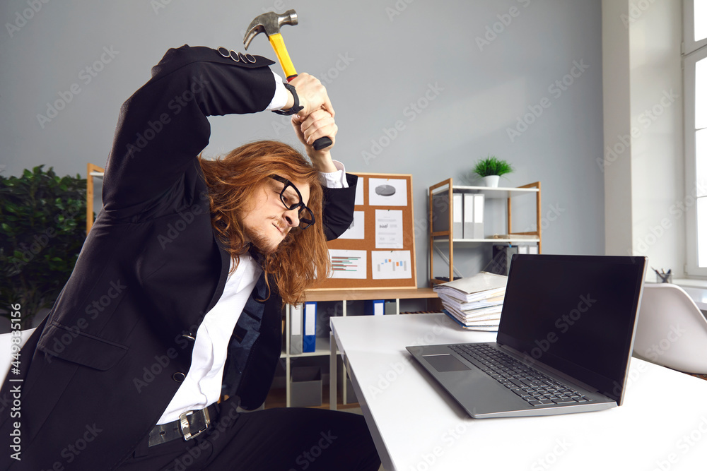 Foto Stock Funny mad young office worker who hates his job having terrible  day. Angry stressed crazy guy hitting laptop with hammer and breaking it  down in rage. Computer failure, error, stress