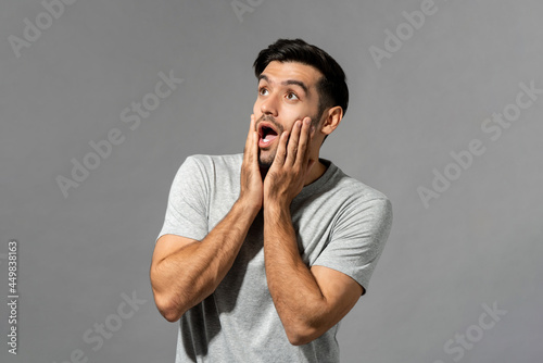 Shocked young Caucasian man with hands on cheeks gasping and looking up in light gray isolated studio background