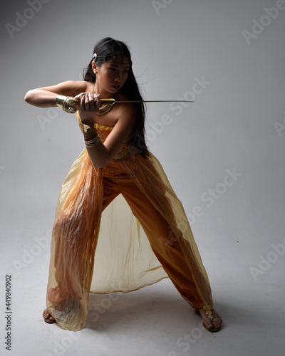 Full length portrait of pretty young asian woman wearing golden Arabian robes like a genie, holding a sword weapon, isolated on studio background. photo