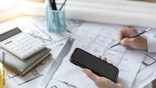 Young man using Smartphone plotting a system of building structures in blueprints, Architects or engineers are designing buildings using mobile to calculate the physical structure to be correct.
