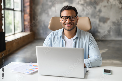 Cheerful indian male student or office worker is using laptop computer sitting at the desk. Friendly and optimistic eastern dark-haired freelancer guy in eyeglasses looks at camera and smiles