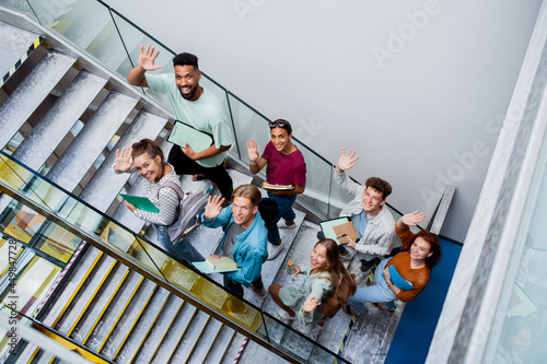 Top view of university students walking up the stairs indoors, looking at camera and waving.