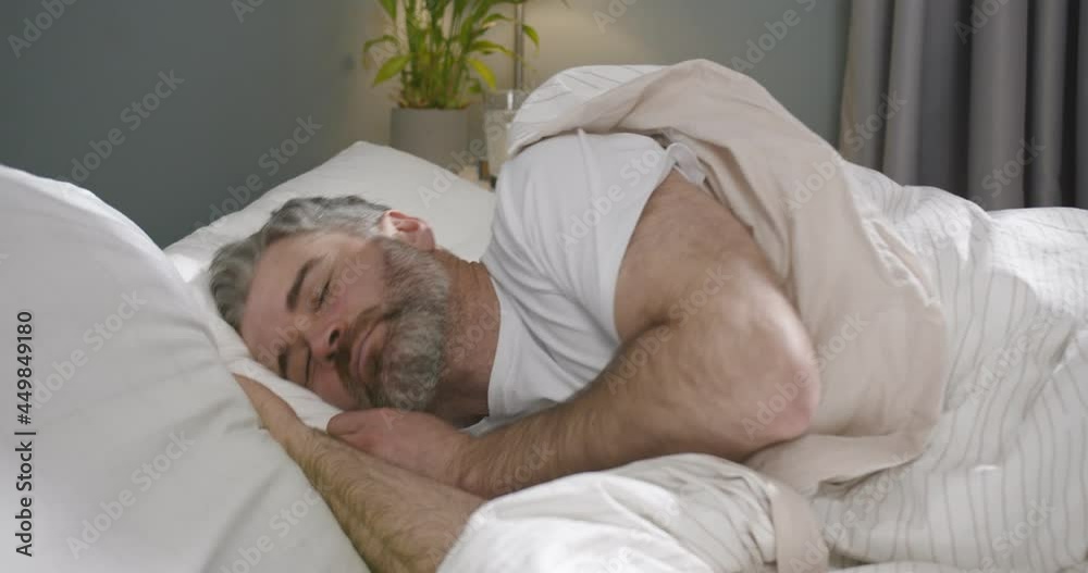 Caucasian handsome middle-aged man with grey beard lying in bed under blanket with eyes closed and sleeping in the morning. Male turning fom side to side in bedroom when sleep at daytime. Caucasian handsome middle-aged man with grey beard lying in bed under blanket with eyes closed and sleeping in the morning. Male turning fom side to side in bedroom when sleep at daytime.