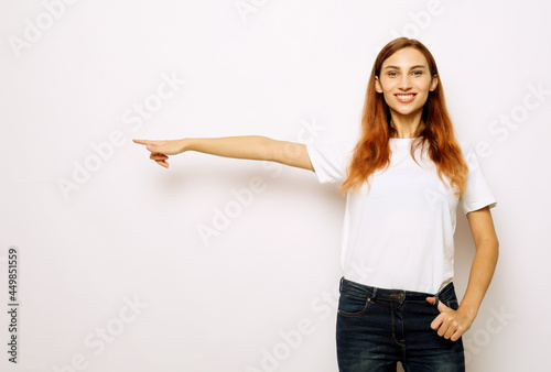 Smiling young red-haired woman isolated on gray studio background pointing with finger at blank copy space aside