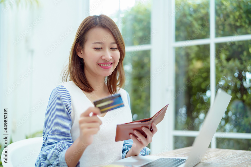 A beautiful Asian woman wearing a white shirt with a credit card comes out of her wallet smiling happily in front of her laptop preparing for online shopping. A beautiful Asian woman wearing a white shirt with a credit card comes out of her wallet smiling happily in front of her laptop preparing for online shopping.