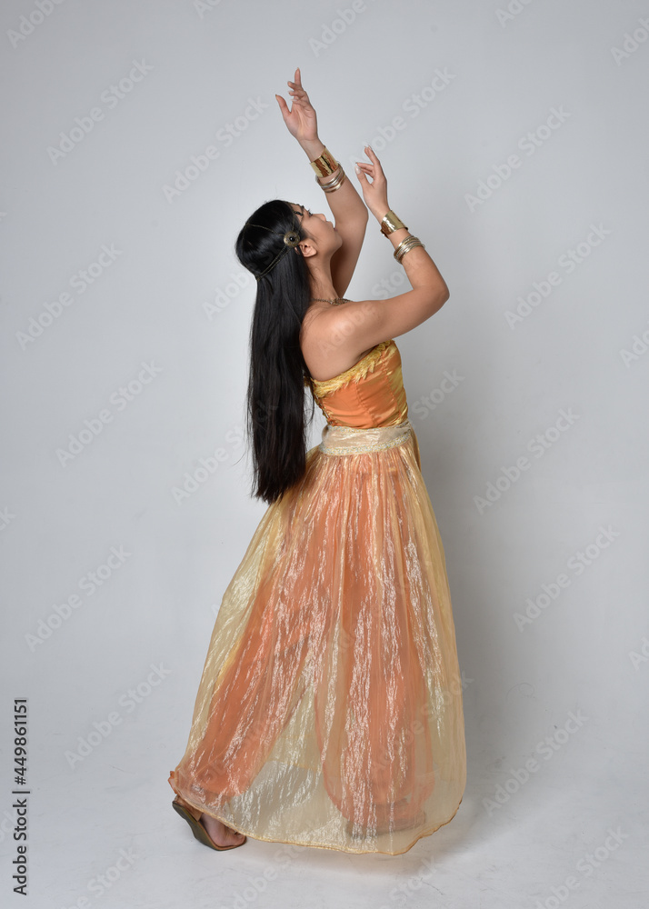 Fototapeta premium Full length portrait of pretty young asian woman wearing golden Arabian robes like a genie, standing pose with back to the camera, isolated on studio background.
