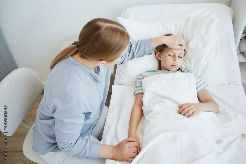 High angle portrait of caring mother by bed of sick child in hospital room, focus on little girl with oxygen support system