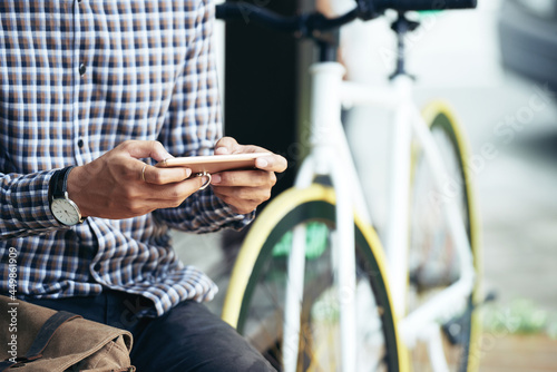 Close-up image of young man sitting next to his bicycle and texting friends or using new popular application on smartphone