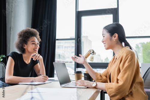 Obraz na plátně asian businesswoman talking to smiling african american colleague near laptop wi