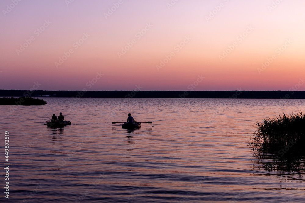 fishermen on boats swim to the shore against the background of a pink-lilac sunset on the lake