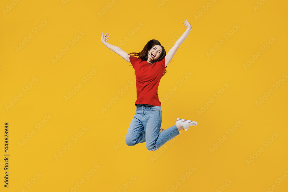 Full size body length excited overjoyed happy young brunette woman 20s wears basic red t-shirt jumping spreading hands isolated on yellow background studio portrait. People emotions lifestyle concept.