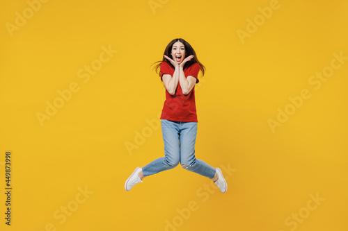 Full size body length shocked surprised fun young brunette woman 20s wears basic red t-shirt jump keep mouth wide open isolated on yellow background studio portrait. People emotions lifestyle concept
