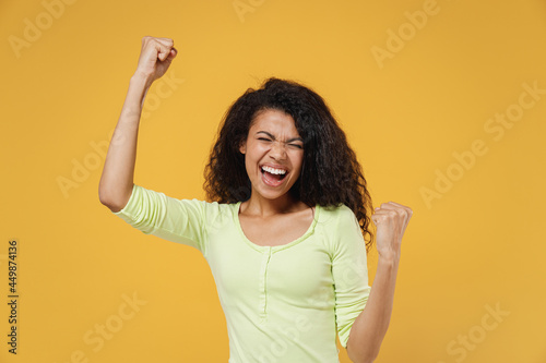 Excited fun african american young woman 20s wears green shirt do winner gesture celebrating clenching fists say yes isolated on yellow background studio portrait. People emotions lifestyle concept