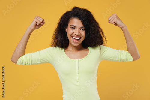 Strong sporty fitness smiling fun african american young brunette woman 20s wears green shirt showing biceps muscles on hand demonstrating strength power isolated on yellow background studio portrait