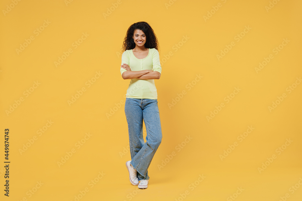 Full size body length smiling vivid happy african american young woman 20s wears green shirt stand hold hands crossed isolated on yellow background studio portrait. People emotions lifestyle concept Full size body length smiling vivid happy african american young woman 20s wears green shirt stand hold hands crossed isolated on yellow background studio portrait. People emotions lifestyle concept